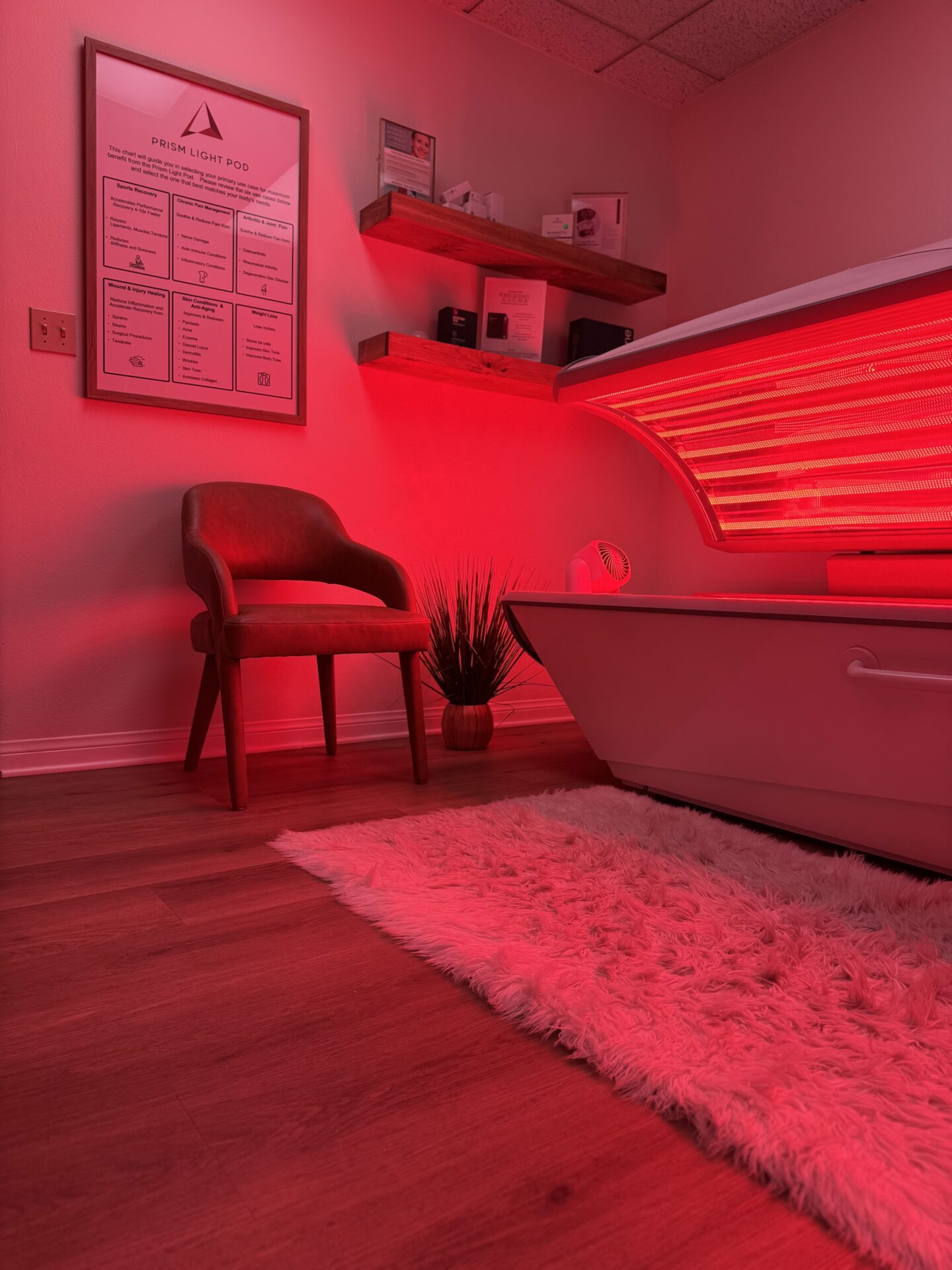 A cozy spa room with light blue brick walls. A massage table with white sheets stands near a wooden console table with decorative baskets and a clock. Two wooden shelves with various products and decor are on the wall. A beige chair with a blue cushion is beside the table.